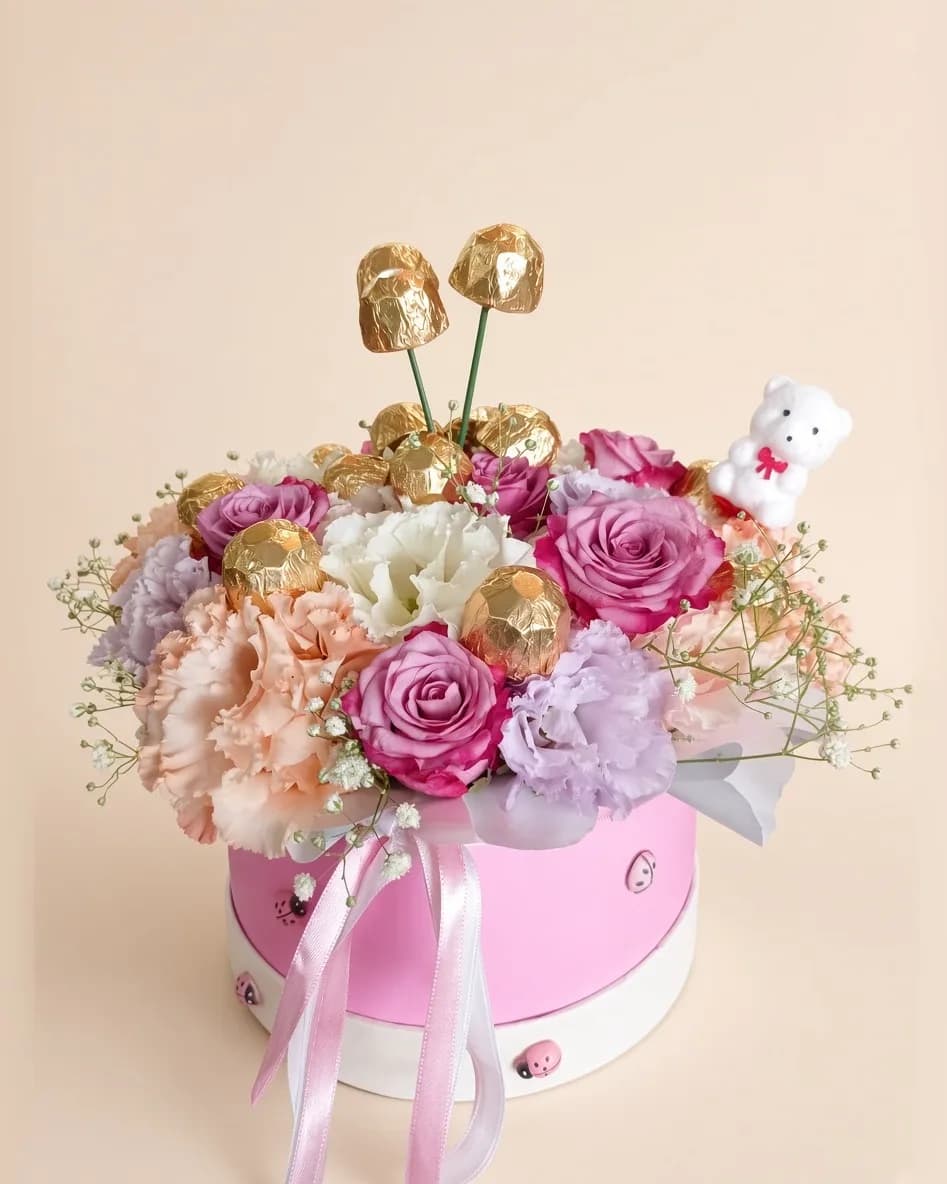 Pembe şapka kutusunda pembe güller, şeftali karanfiller, altın ambalajlı çikolatalar ve beyaz peluş ayıcık